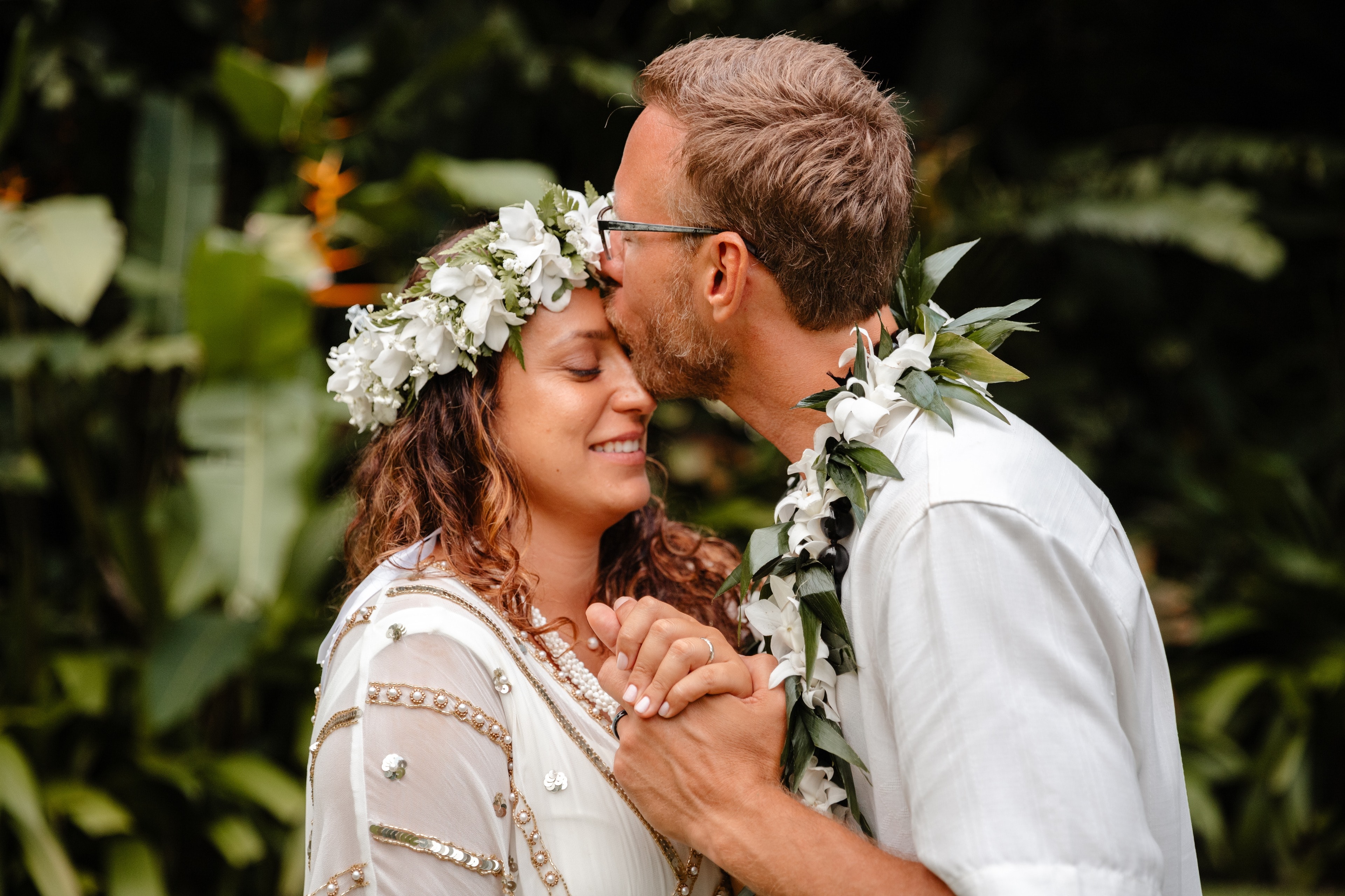 A groom kissing the bride's forehead, both wearing flower lei